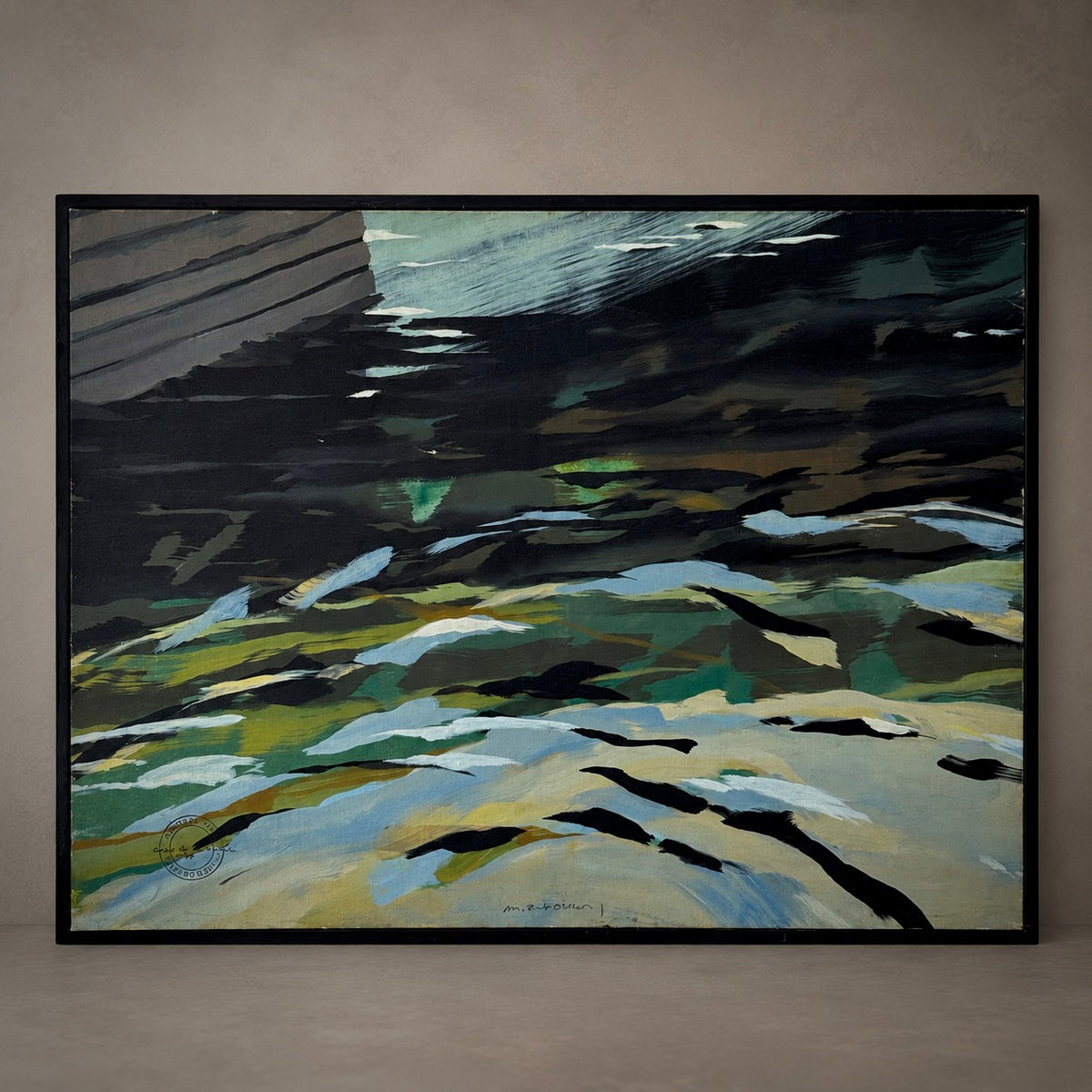 An abstract framed painting depicting a textured landscape with dark black and gray areas, green and blue-green patches, and sandy beige tones, hung on a neutral gray wall.