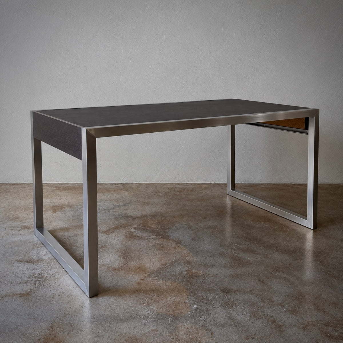 A modern rectangular desk with a dark gray wood-grain top and a brushed silver metal frame, set against a neutral gray wall and concrete floor.