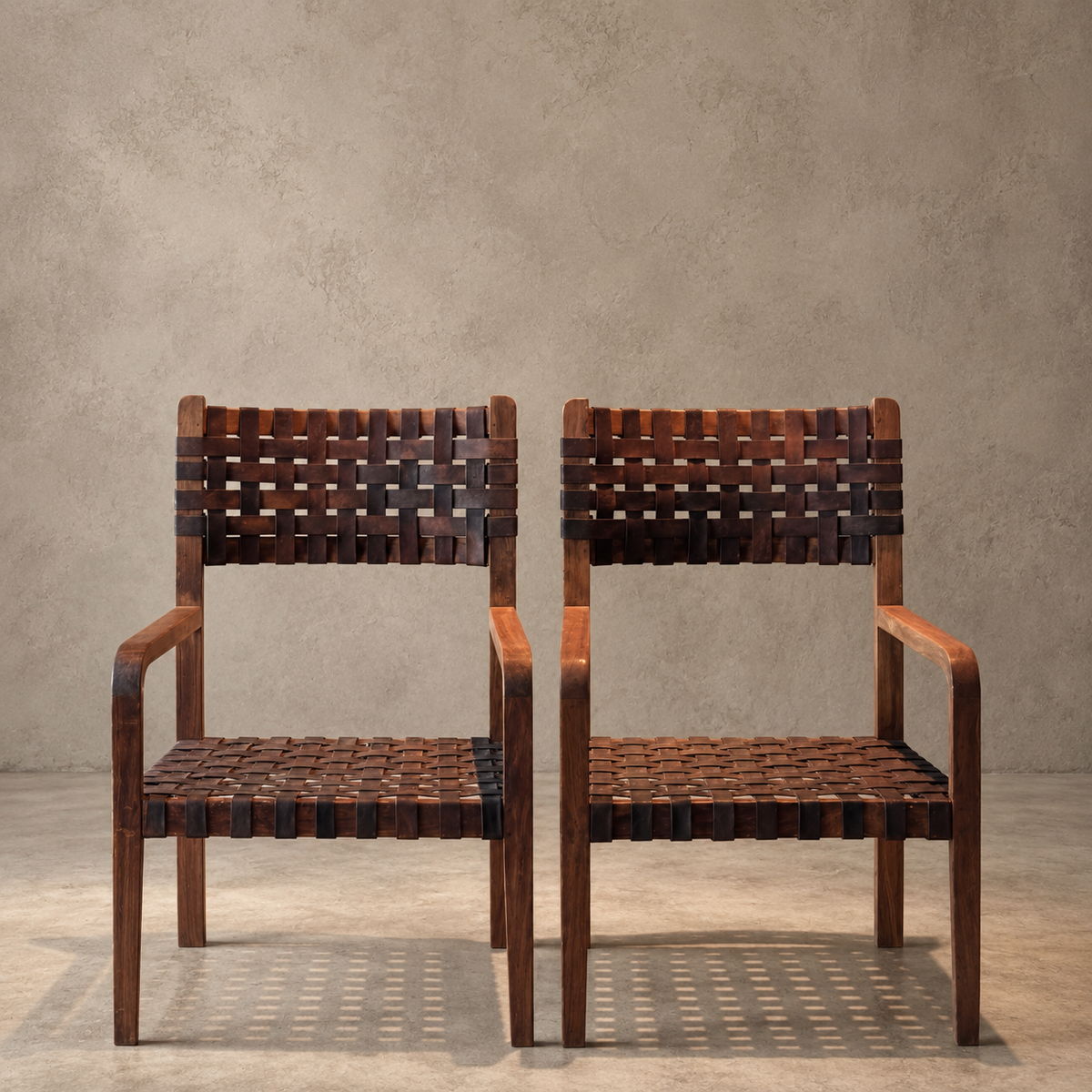 Two wooden armchairs with woven dark brown and black leather seats and backrests, set against a textured gray concrete wall and floor.