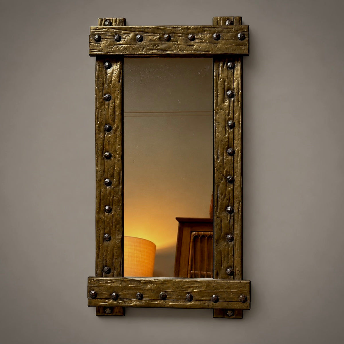A rustic rectangular wall mirror with a thick, distressed bronze-brown wooden frame featuring dark metal rivets, shown against a neutral gray wall.