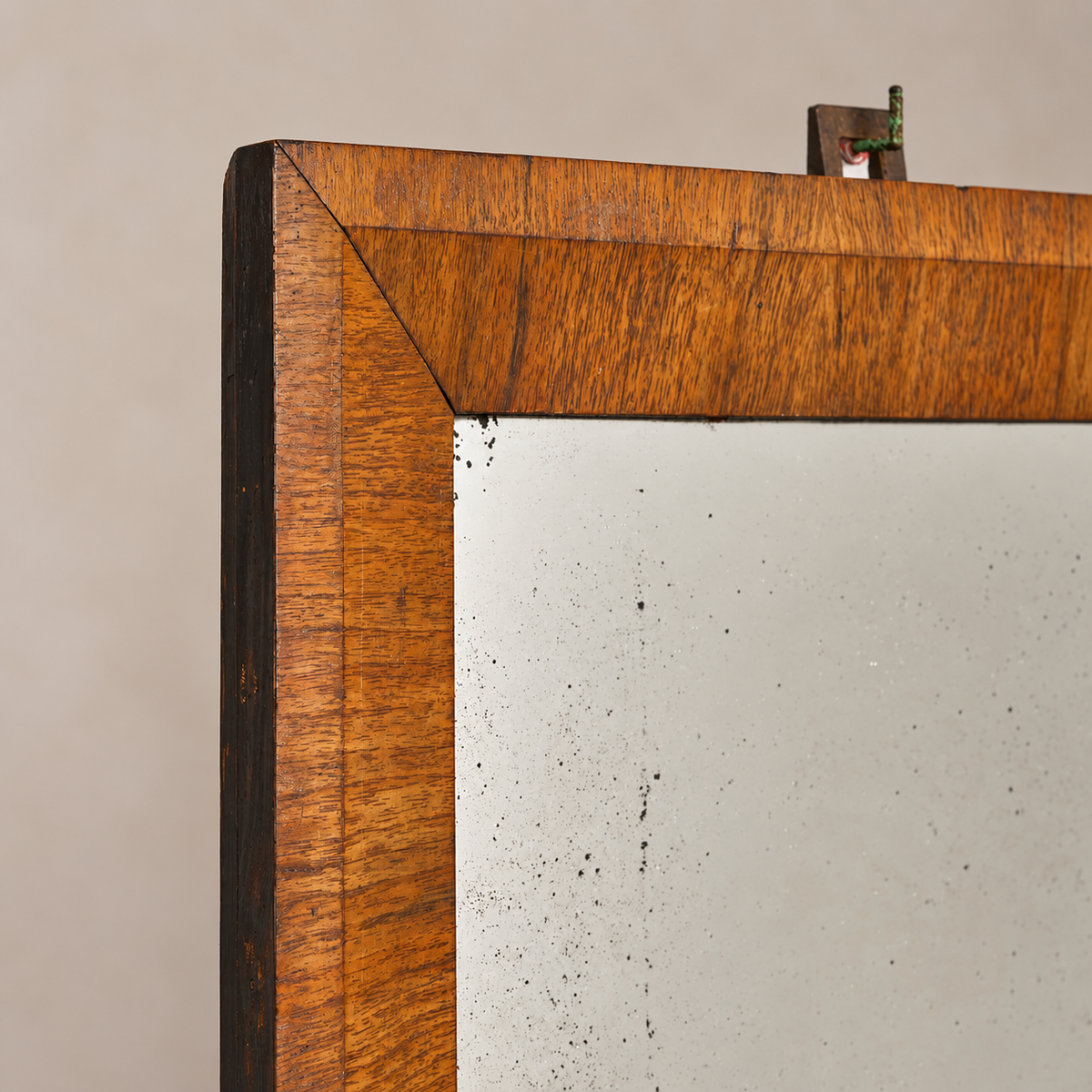 Close-up of a wooden-framed mirror corner, showing a warm medium-brown wood grain border and a light off-white mirror surface against a plain beige wall.