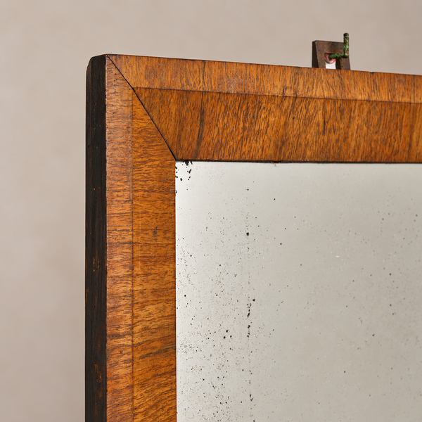 Close-up of a wooden-framed mirror corner, showing a warm medium-brown wood grain border and a light off-white mirror surface against a plain beige wall.