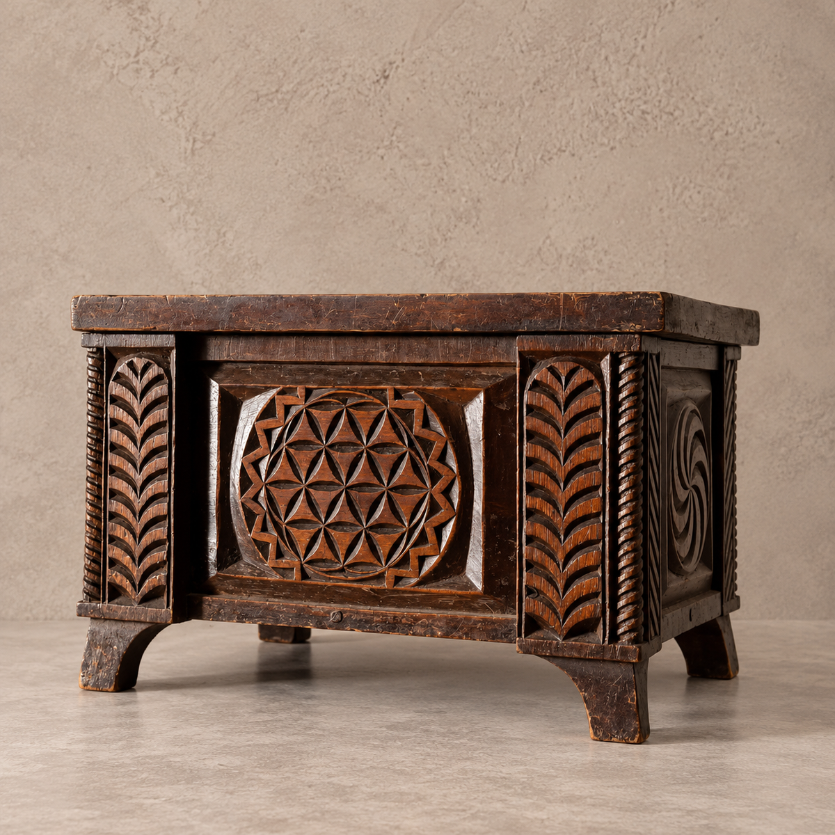 A dark brown carved wooden box with ornate geometric and leaf-like relief patterns on the front and sides, photographed against a textured beige wall and light floor.