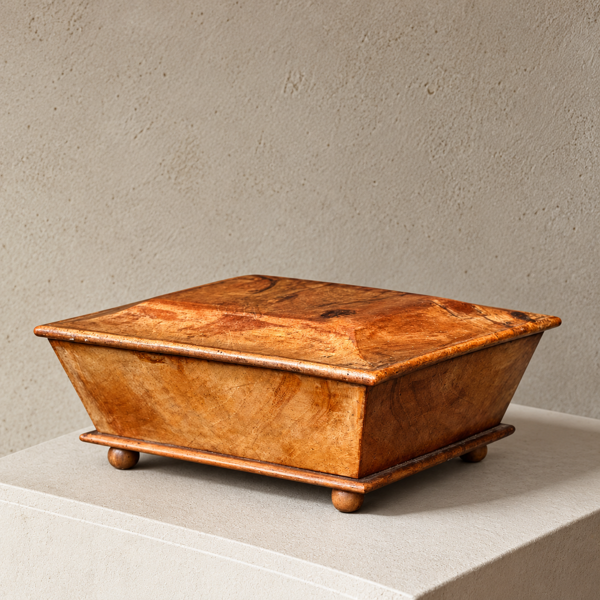 A rectangular wooden box with a hinged lid, shown closed on a light stone pedestal against a textured beige wall. The wood has a warm honey-brown finish with visible grain patterns and small round feet.