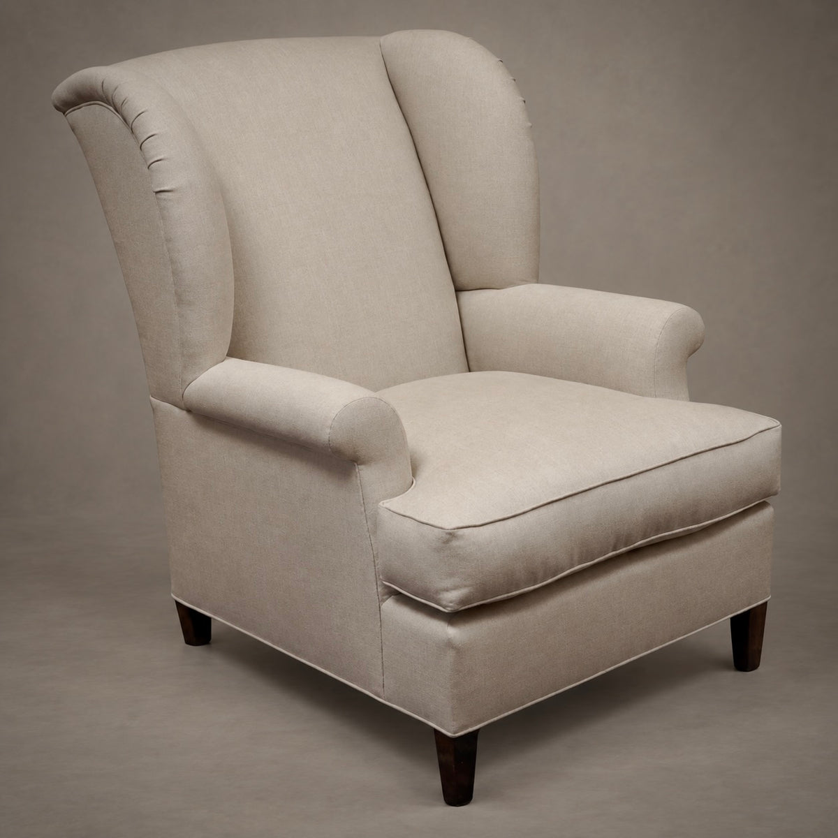 An upholstered wingback armchair in a light beige fabric, featuring dark wooden legs, photographed against a neutral gray studio background.