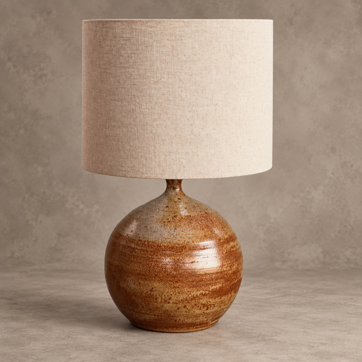 A table lamp with a round, glossy brown ceramic base and a large, cylindrical beige linen drum shade, set against a textured neutral gray background.