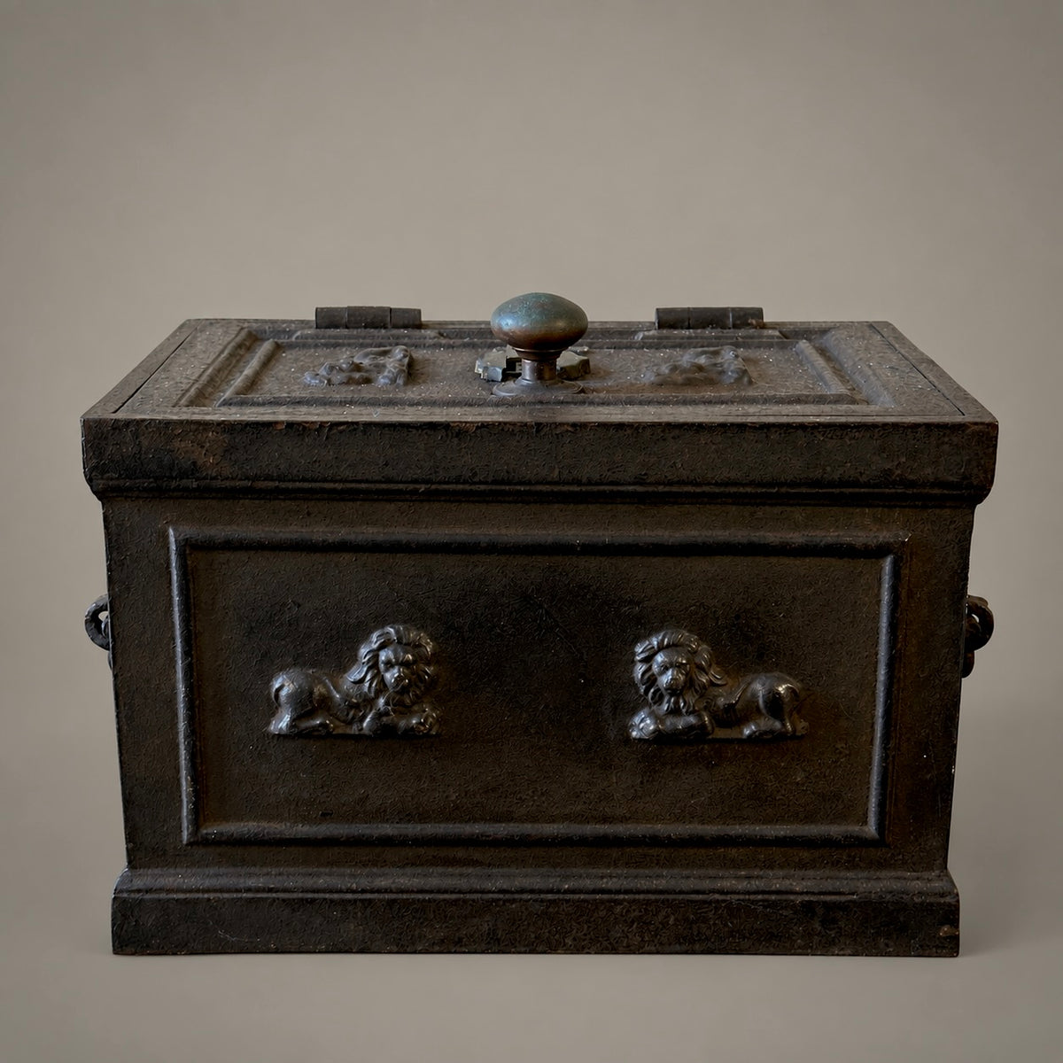 An antique dark bronze or black metal chest with lion head handles on the front and a round knob on top, photographed against a plain beige background.