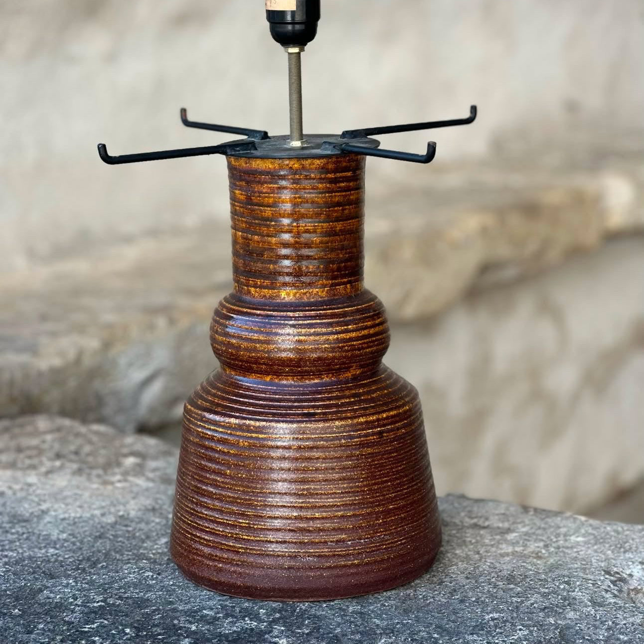 A rustic brown ceramic table lamp with a ribbed textured base, a black metal cross-shaped shade holder, and a clear glass bulb in a black socket. The lamp is displayed outdoors on a stone surface with a blurred stone wall background, and text overlay on the right shows dimensions and price.
