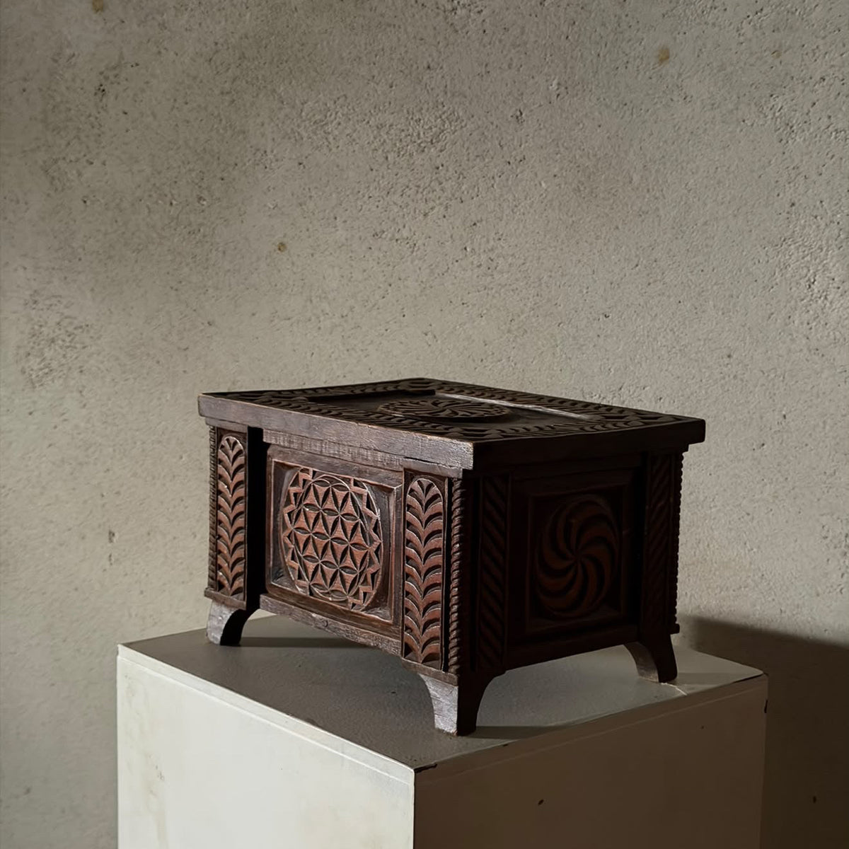 An ornate dark brown carved wooden chest with geometric and floral patterns, displayed on a white pedestal against a textured concrete wall.