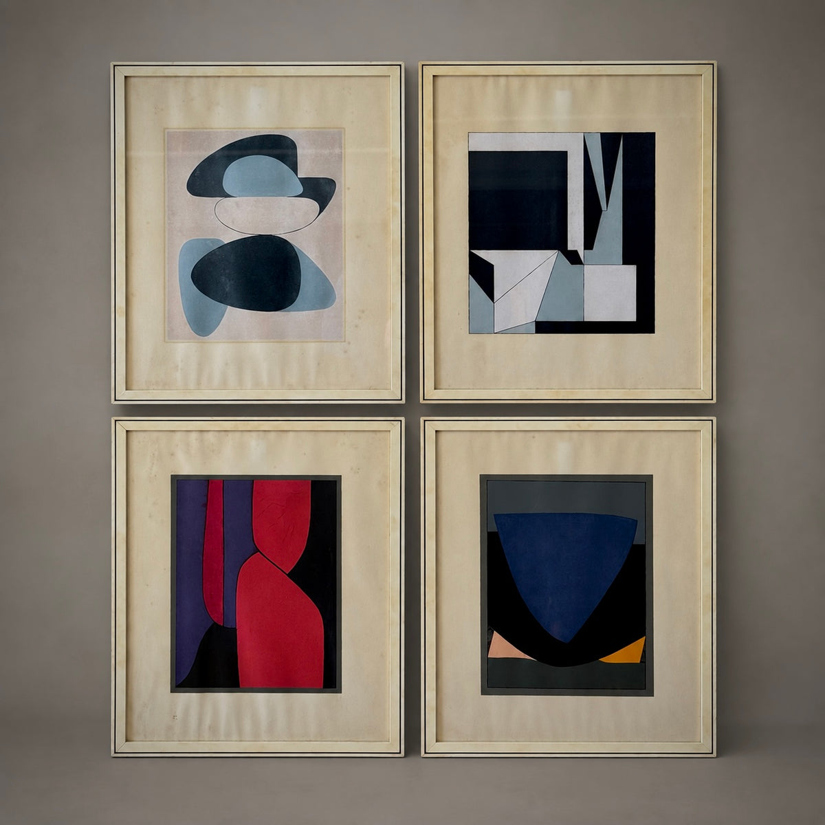 Four framed abstract geometric art prints arranged in a 2x2 grid on a neutral gray wall. The frames are light beige wood, and the artworks feature a mix of navy, blue, red, black, and white shapes.