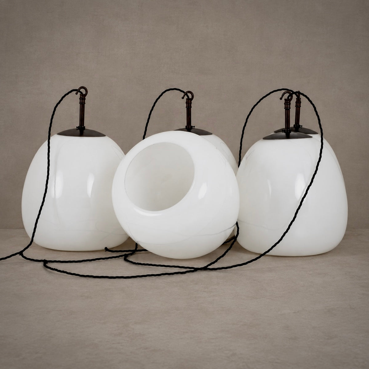 Three white teardrop-shaped pendant light shades with black hanging cords and tops, arranged on a neutral beige studio surface and backdrop.