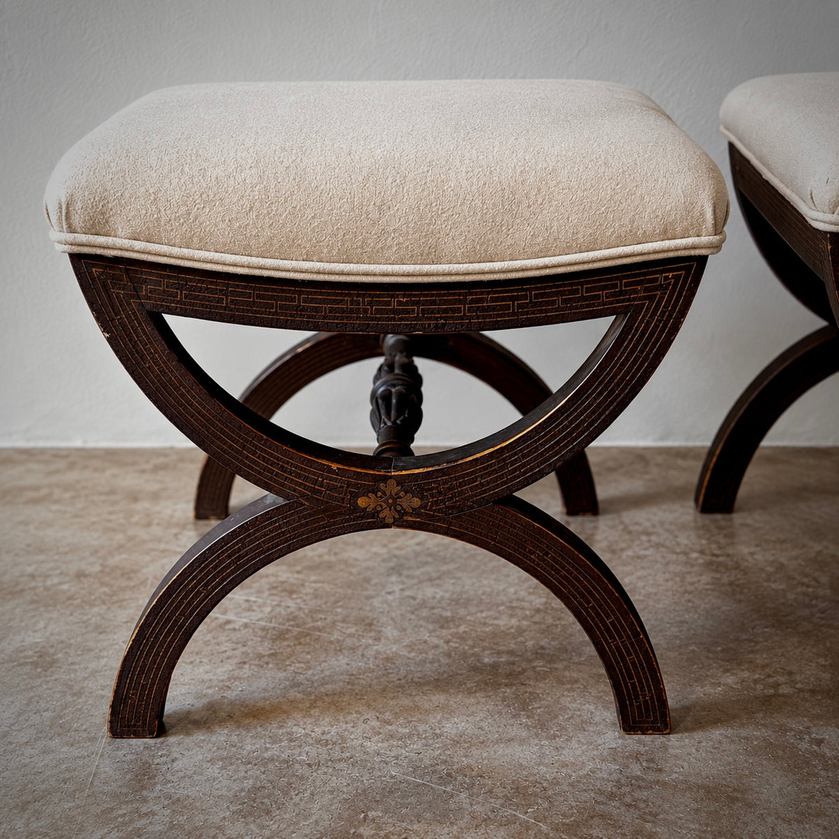 A dark brown wooden stool with a curved, geometric carved frame and a cream-colored upholstered cushion, shown in a minimalist indoor setting.