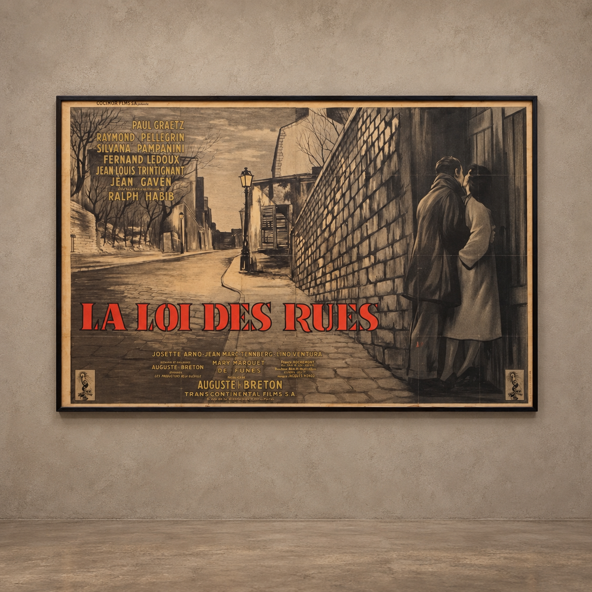 A framed vintage-style movie poster for 'La Loi des Ruines' hanging on a textured gray wall. The poster features a sepia-toned illustration of a street scene with two men standing by a wall, bold red title text, and black credits on a tan background.