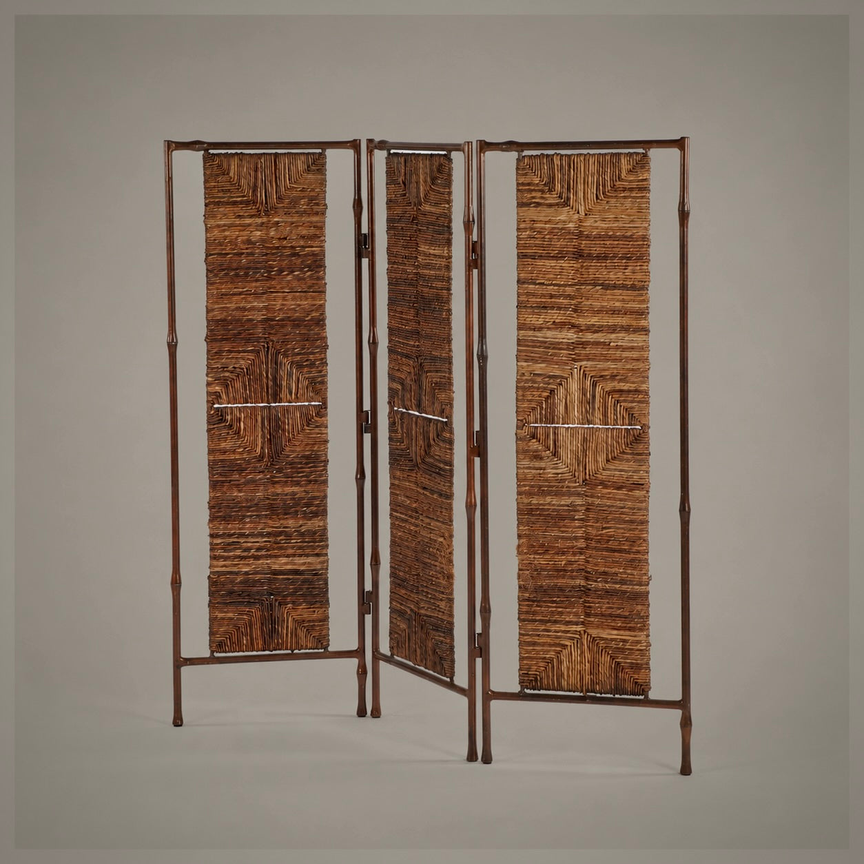A three-panel folding screen with a dark brown bamboo-look metal frame and woven brown raffia or straw panels, shown against a neutral gray studio background.