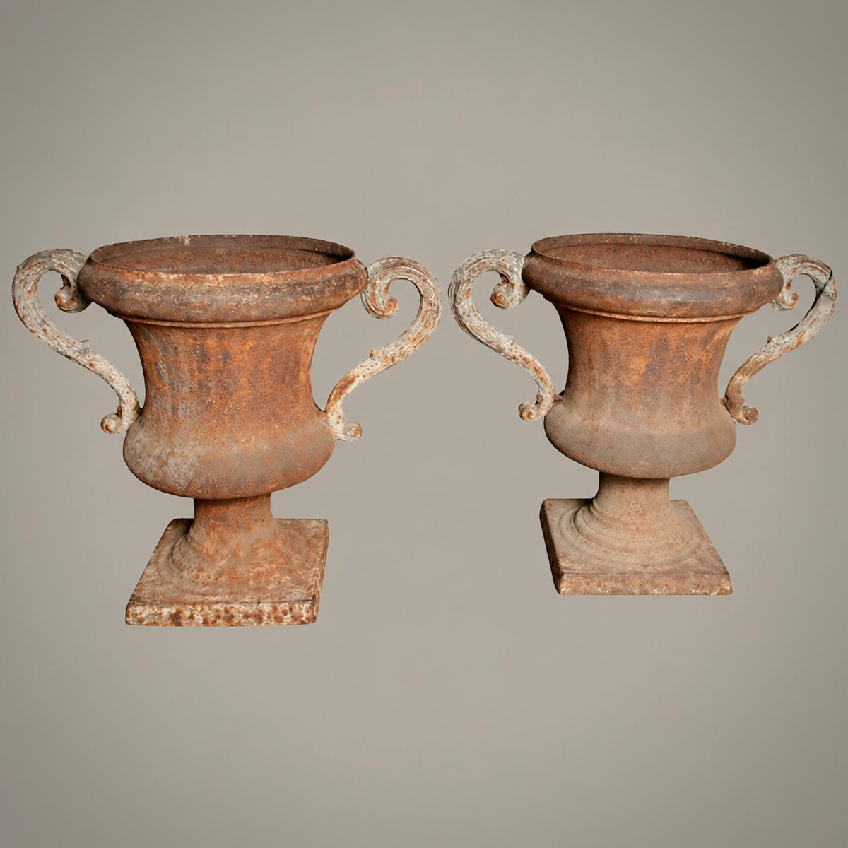 A pair of rustic, weathered terracotta-colored metal urn planters with scrolled handles and square bases, set against a solid neutral gray background.