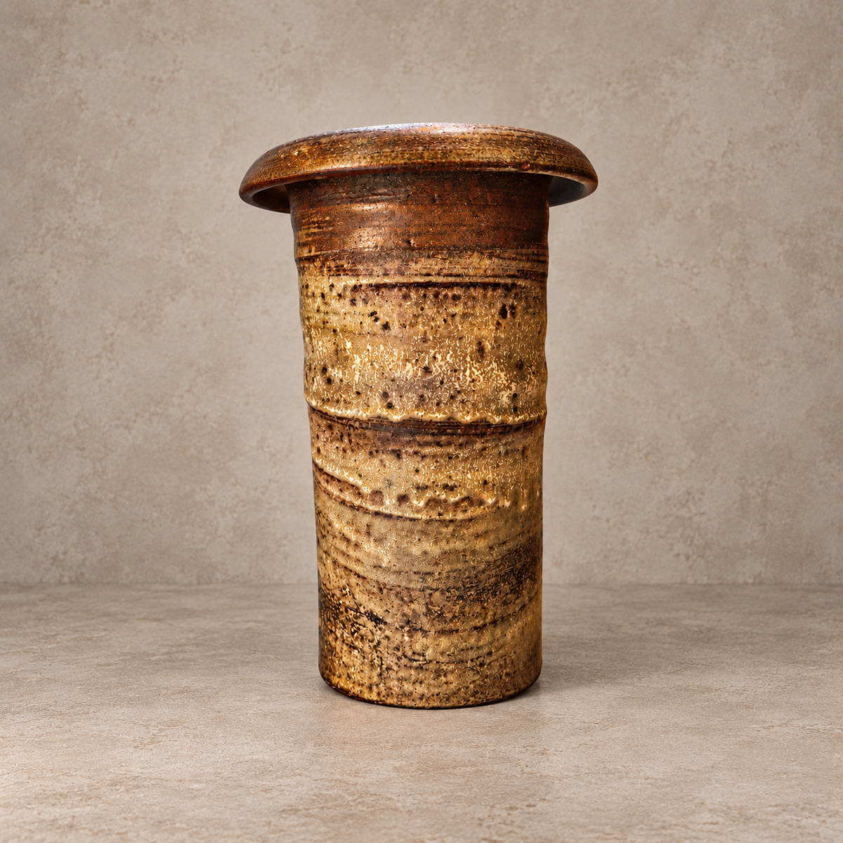 A rustic, cylindrical ceramic pedestal or side table with a wide, flared top, featuring a textured glaze in earthy brown, gold, and beige tones, set against a neutral textured gray background.
