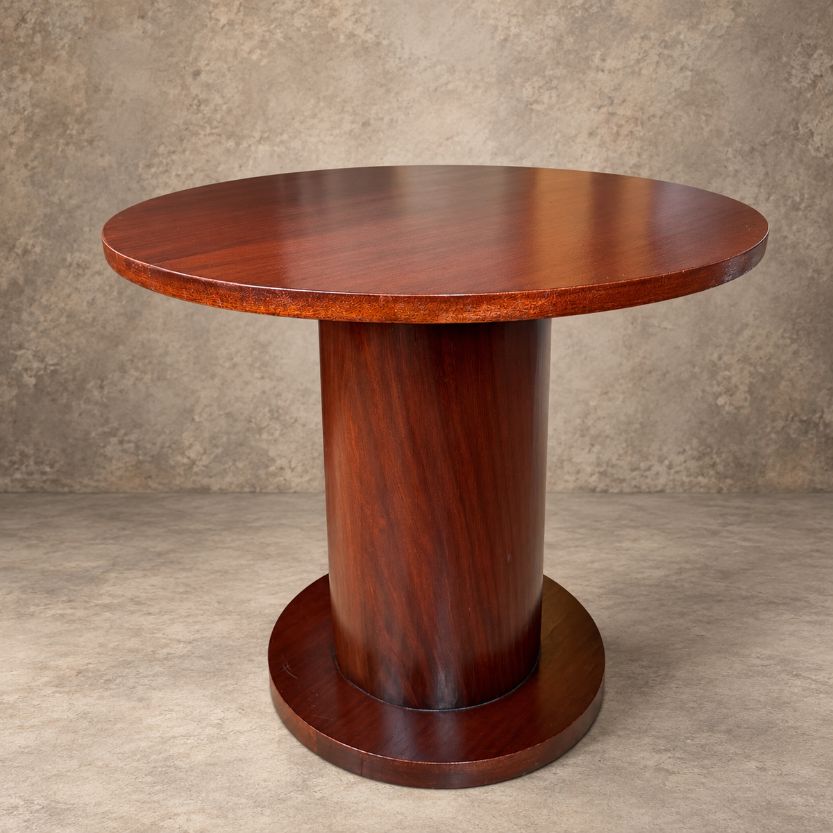 A round wooden pedestal table with a polished mahogany finish, set against a textured concrete wall and floor.