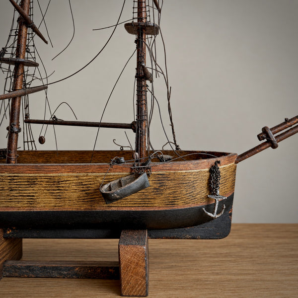 A detailed wooden model of a historic sailing ship with dark brown hulls, weathered wood textures, and a small silver anchor hanging from the side, displayed on a wooden stand against a neutral background.