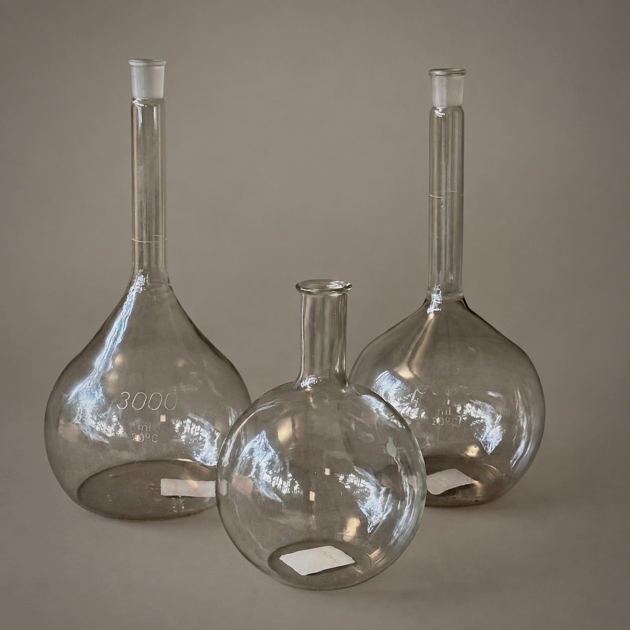 Three clear glass round-bottom flasks with long necks and stoppers, arranged on a neutral taupe background. The front flask is empty, while the two back flasks are partially filled with liquid to demonstrate capacity.
