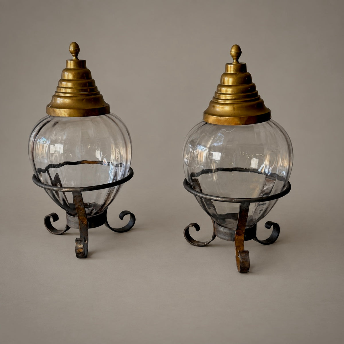 A pair of vintage-style clear glass globe lanterns with ornate brass-toned lids and dark metal scroll stands, photographed against a plain warm gray background.