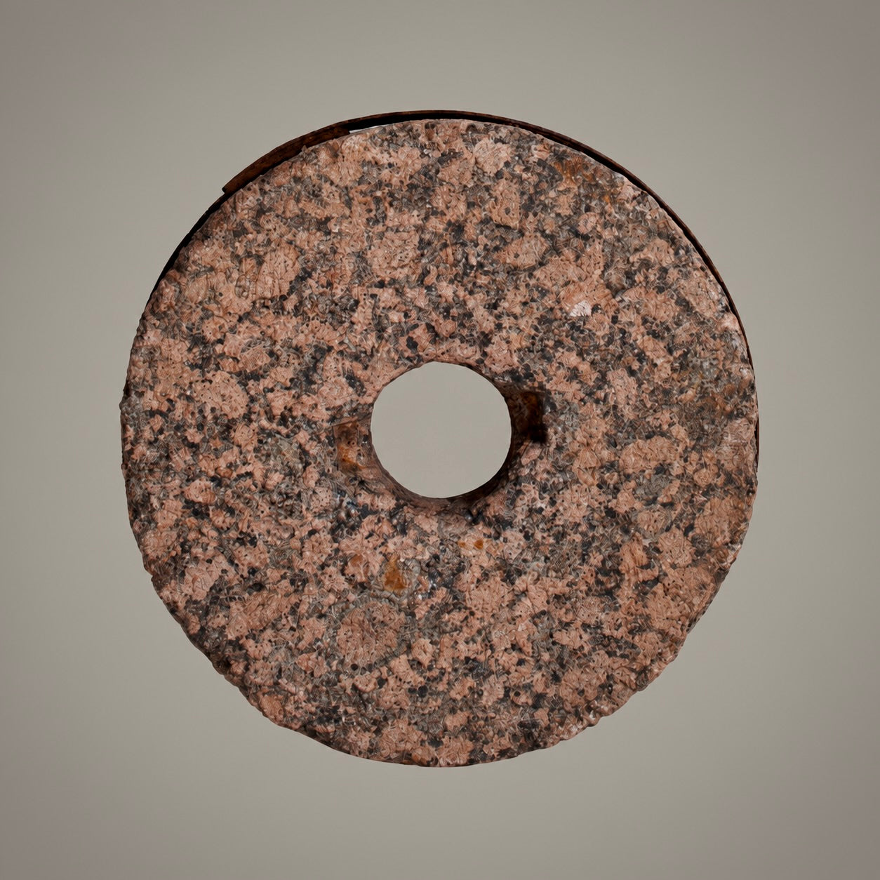 A circular stone or granite ring with a central hole, showing a speckled pink, brown, and black texture, photographed against a solid neutral gray background.