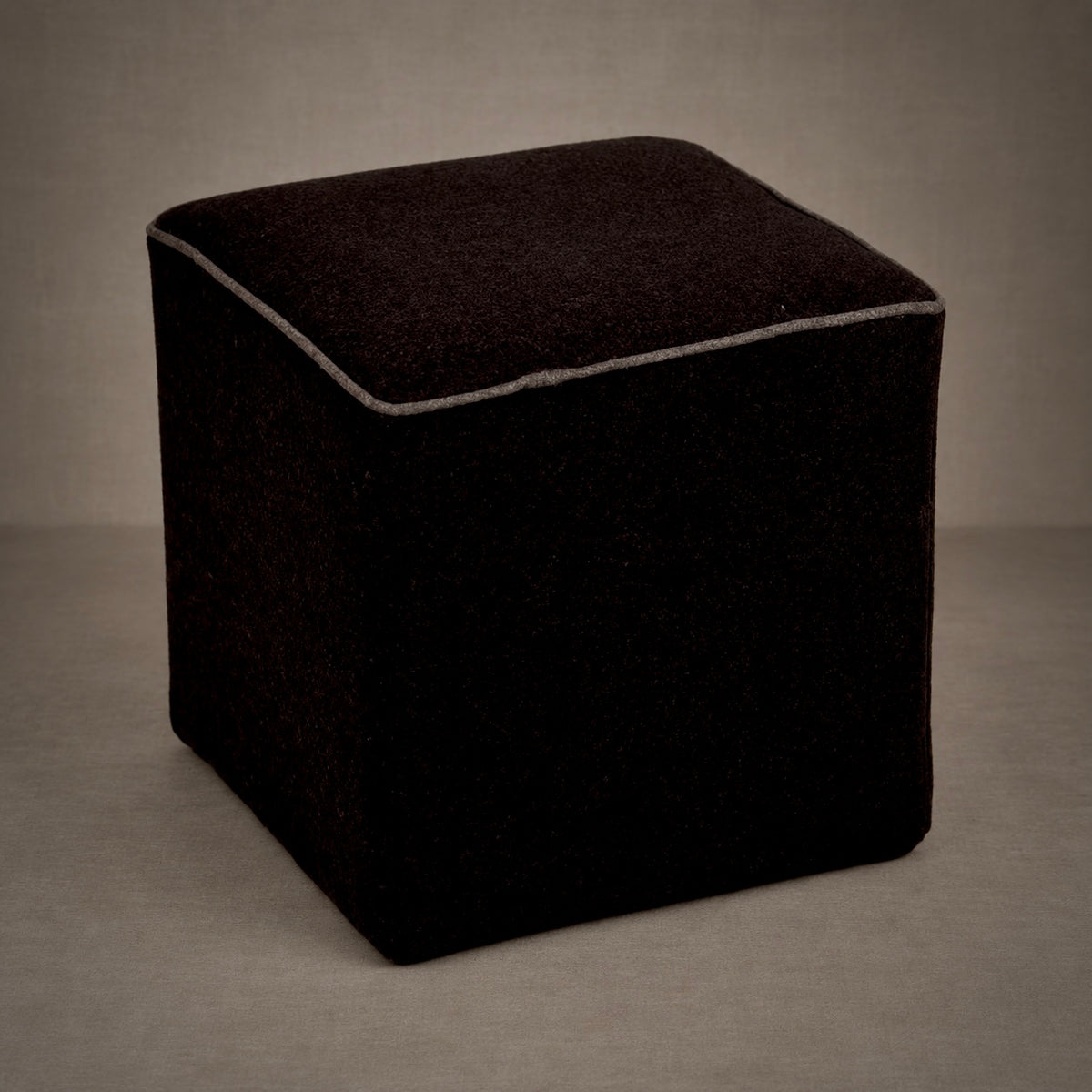 Dark brown upholstered cube ottoman with contrasting light piping, photographed against a neutral tan-gray studio backdrop.