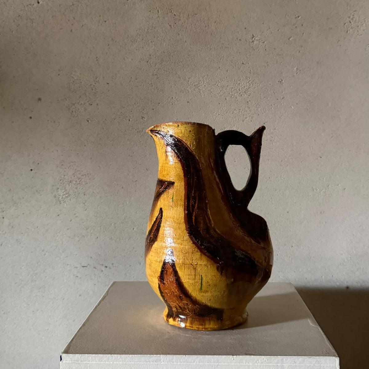 A handmade ceramic pitcher with a glossy finish, featuring a marbled pattern of ochre yellow, brown, and black, displayed on a white pedestal against a textured gray wall.