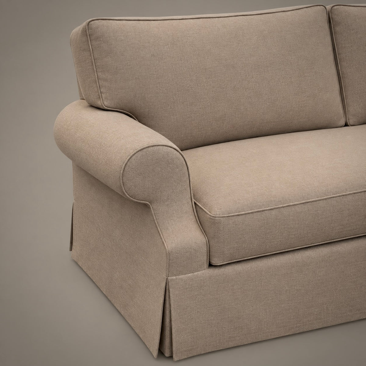 Close-up of a beige upholstered sofa section with rolled arms, a skirted base, and clean tufted seams on a neutral studio background.