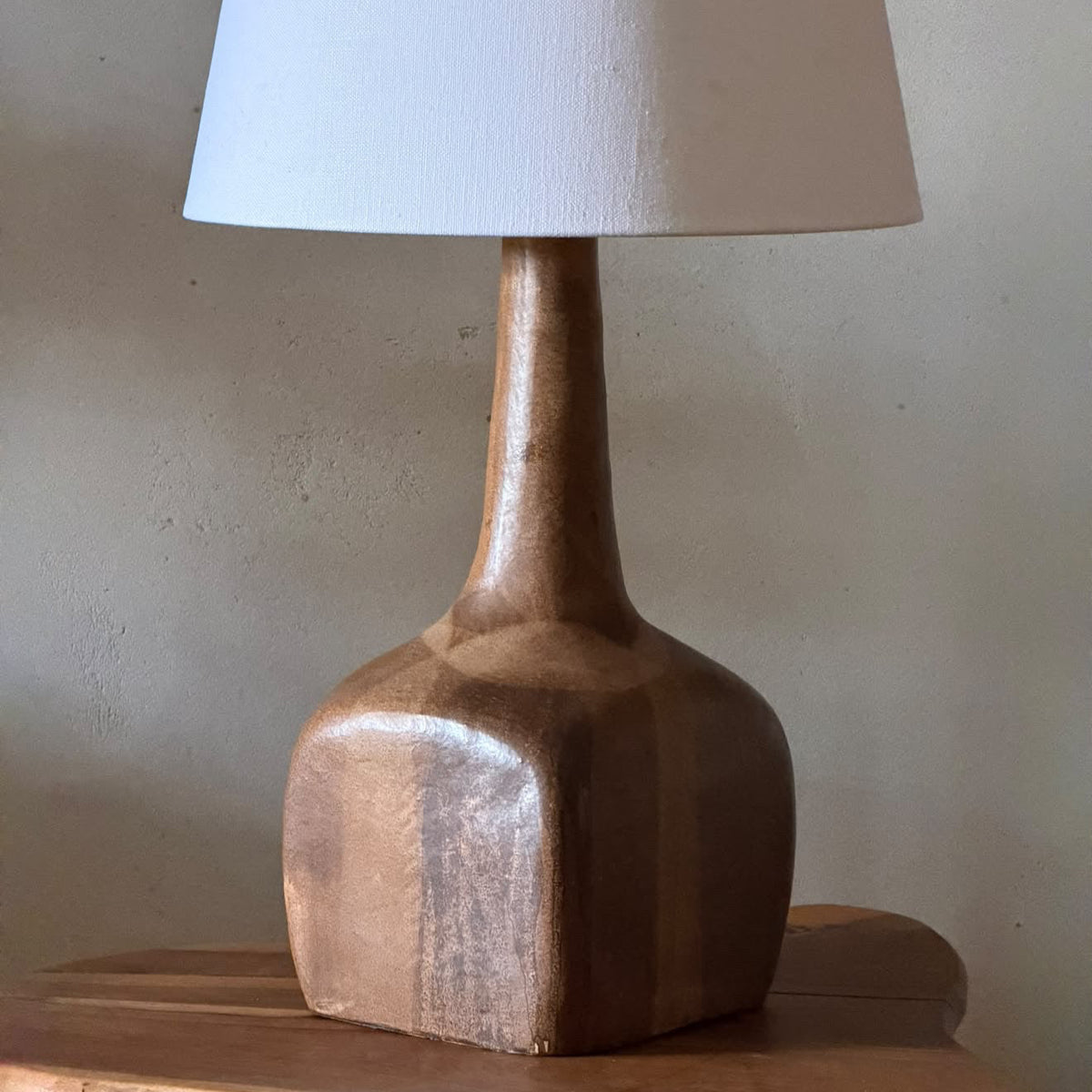 A sculptural table lamp with a tapered white fabric shade and a polished wooden base, displayed on a rustic wooden table against a textured beige wall.