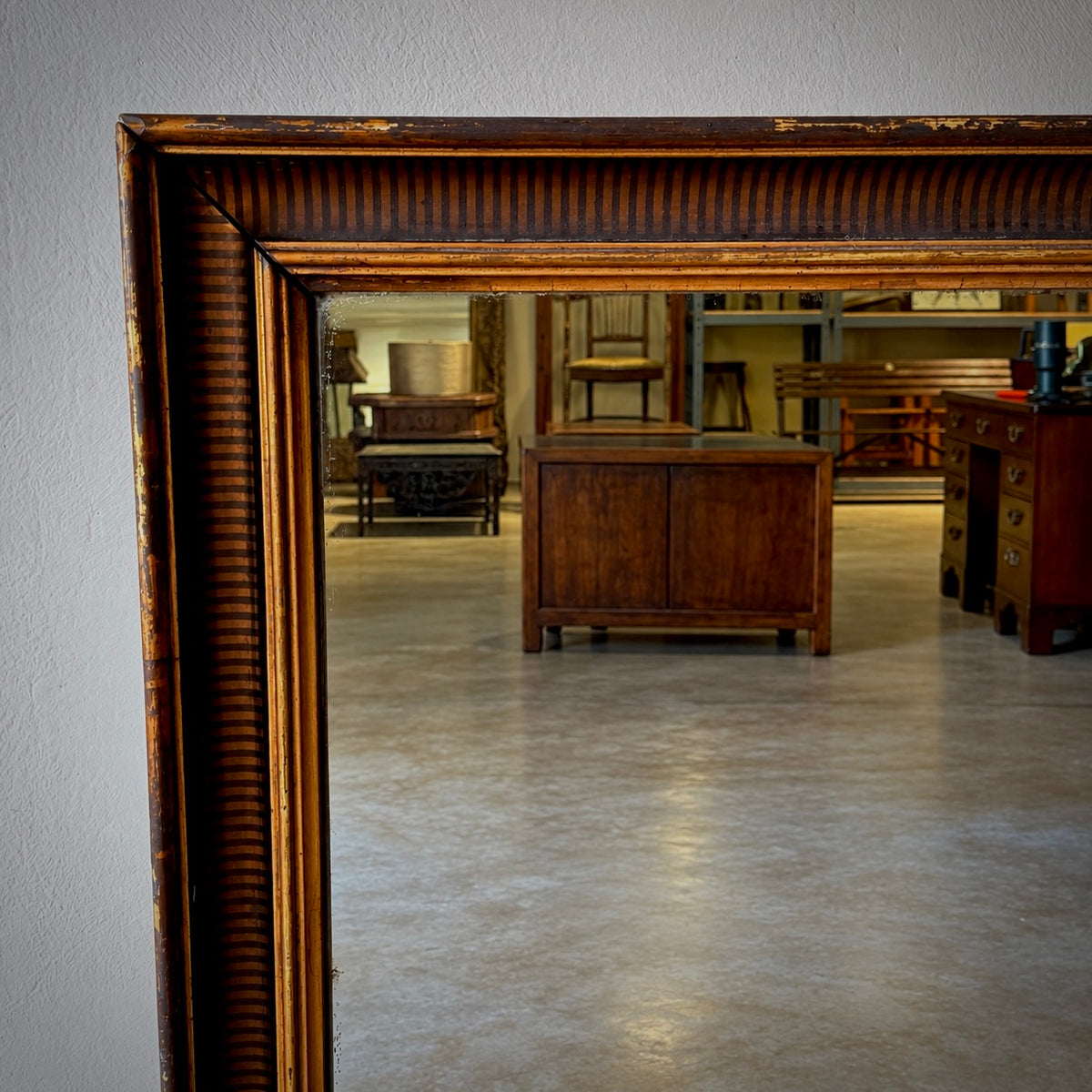 A large antique-style wooden mirror with an ornate, distressed brown frame, reflecting a furniture showroom interior.