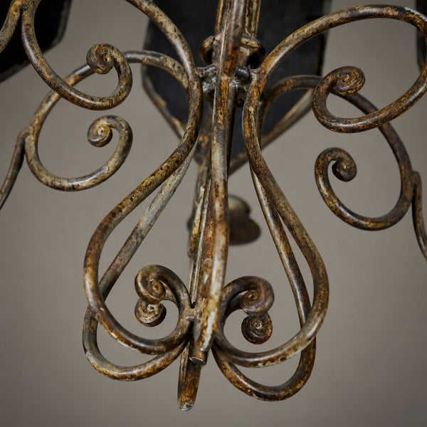 Close-up of an ornate wrought iron chandelier detail, showing scrolled metal arms with a distressed bronze and gold finish against a neutral gray background.