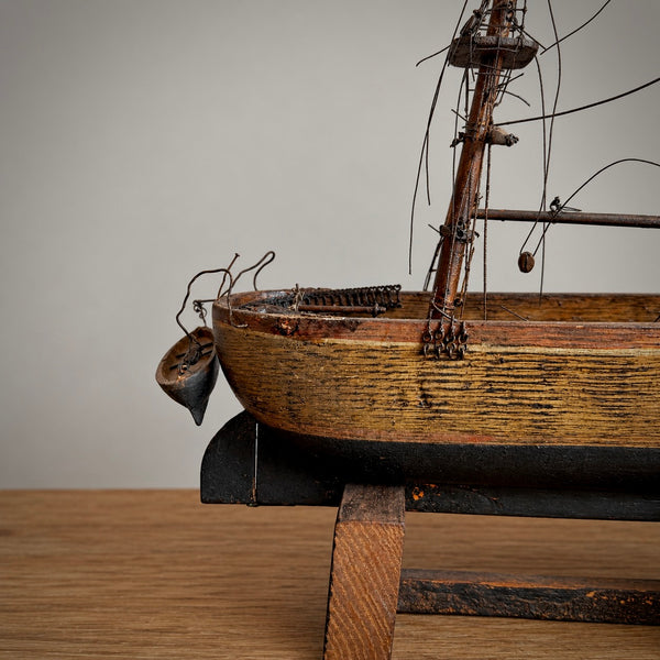 Close-up of a wooden model ship on a black and wood display stand, showing the hull, mast, rigging, and a small hanging weight against a plain light background.