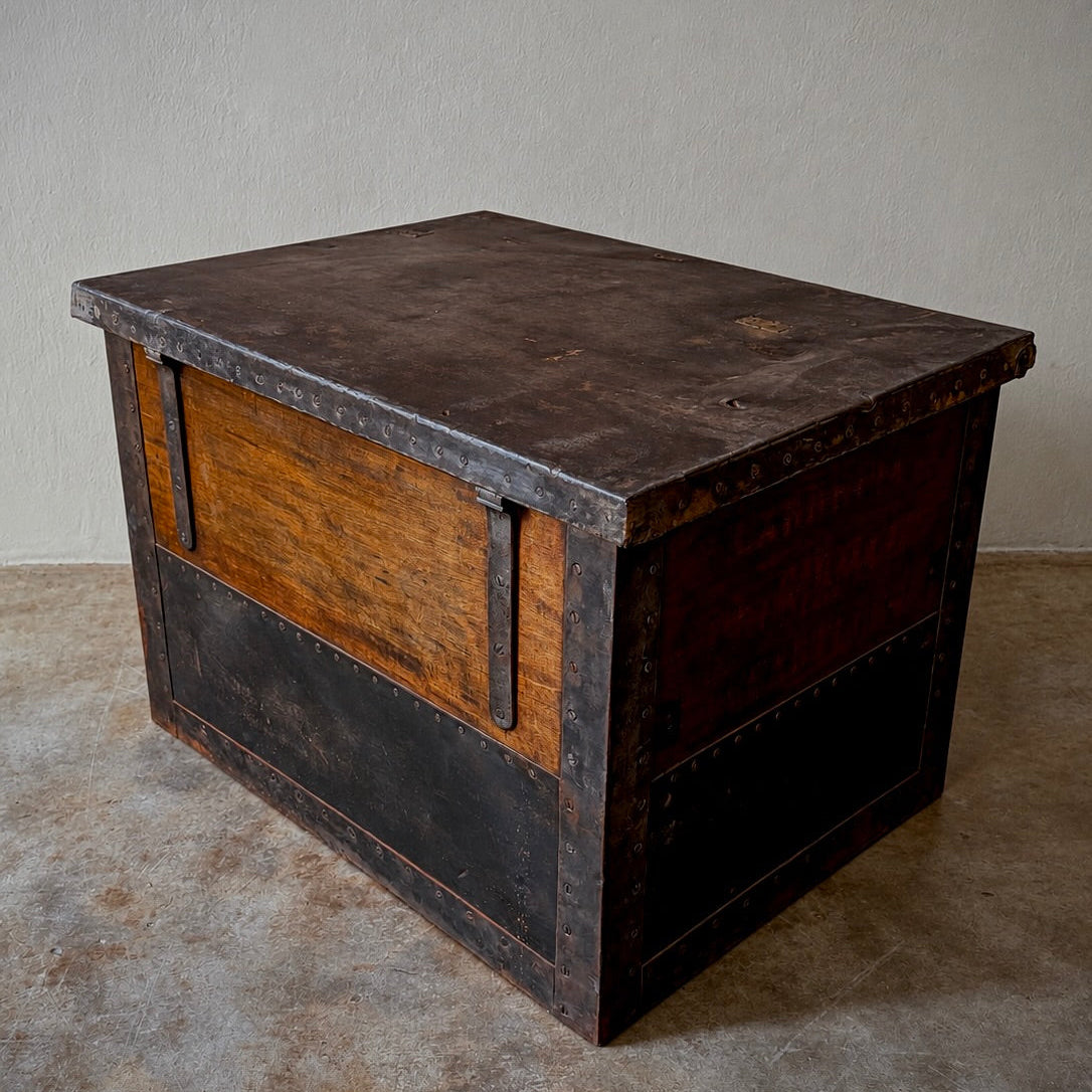 A rustic dark wood chest or trunk with a weathered metal-look top and trim, photographed in a simple indoor room with a plain wall and concrete floor.
