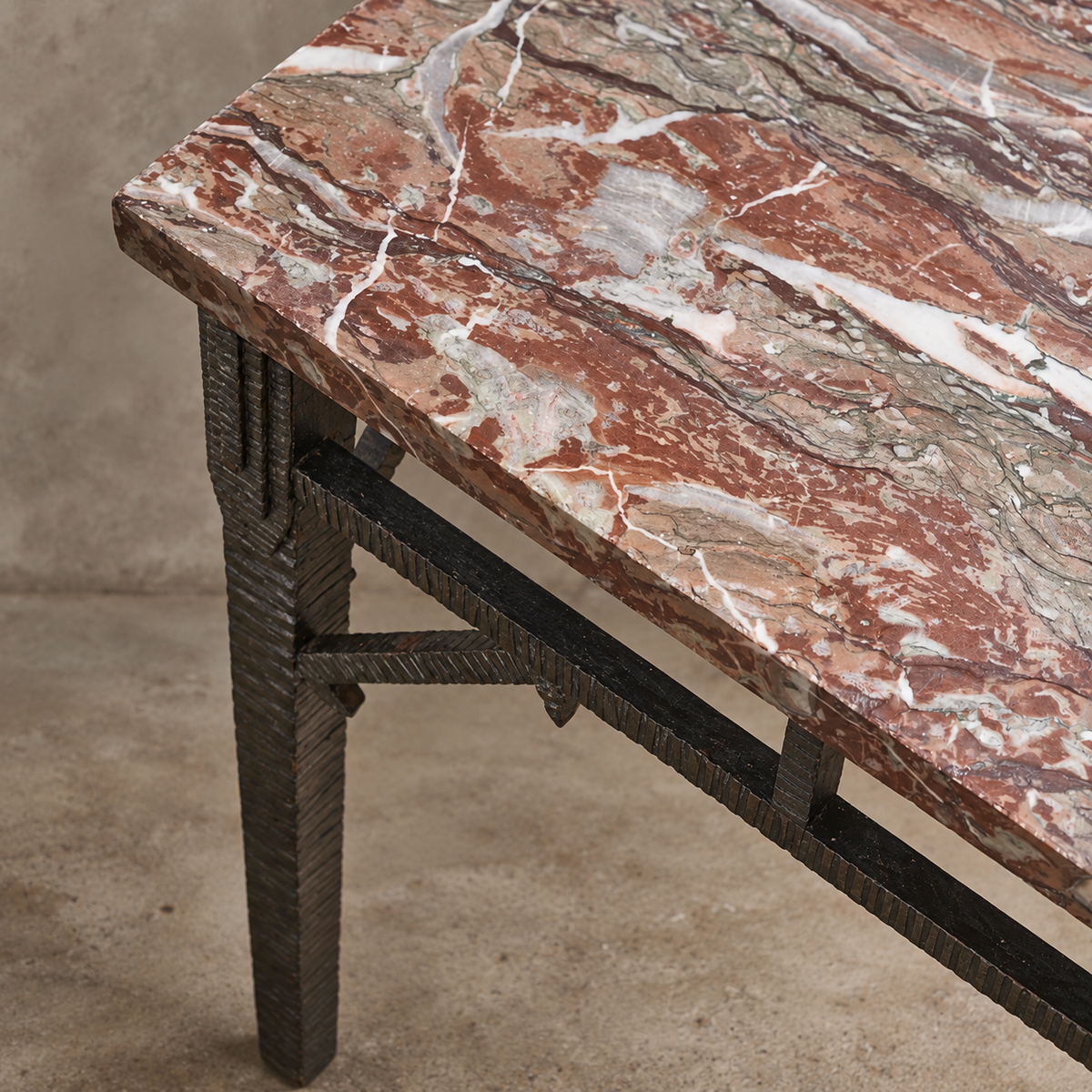 Close-up of a marble-top table with a reddish-brown and cream veined surface and a dark metal frame leg, shown against a concrete floor and wall.