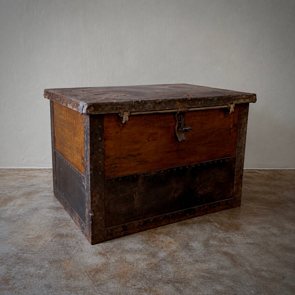 A vintage wooden storage chest with dark metal trim and a latch closure, photographed in a minimalist studio setting with a textured concrete floor and neutral wall.