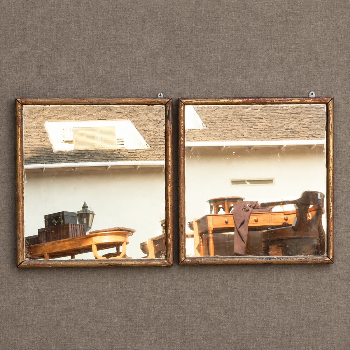 Two rectangular framed mirrors with distressed gold frames mounted side-by-side on a textured gray wall. The mirrors reflect a home interior with wooden furniture, a lamp, and a house exterior roof.
