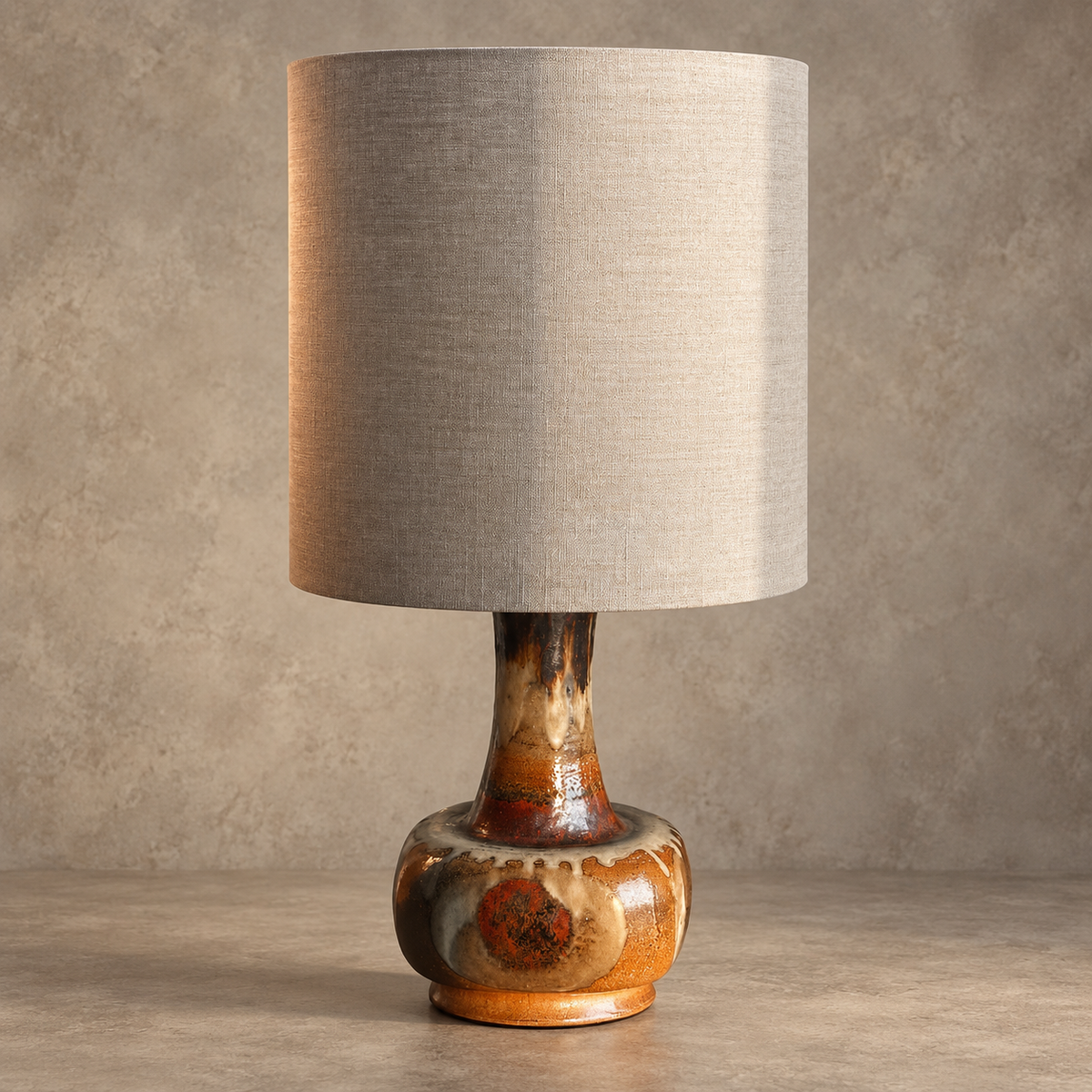 A table lamp with a large beige linen drum shade and a glazed ceramic base in earthy brown, orange, and cream tones, set against a textured neutral background.