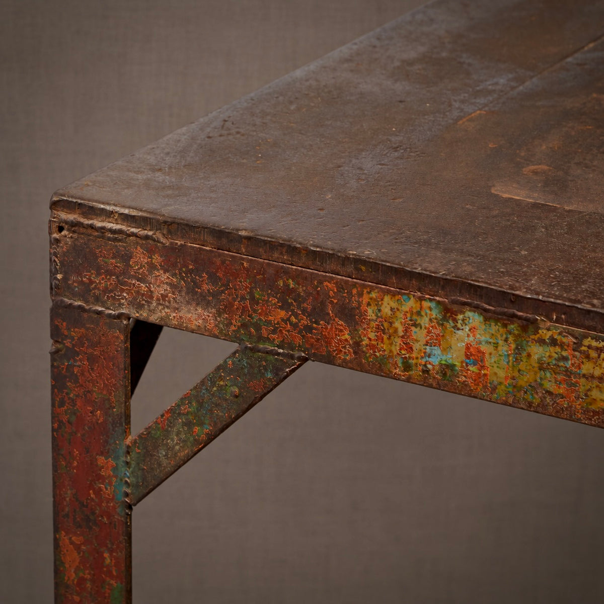 Close-up of a rustic industrial metal table corner with heavily distressed, rusted, and peeling paint showing weathered orange, green, and brown tones against a neutral gray background.