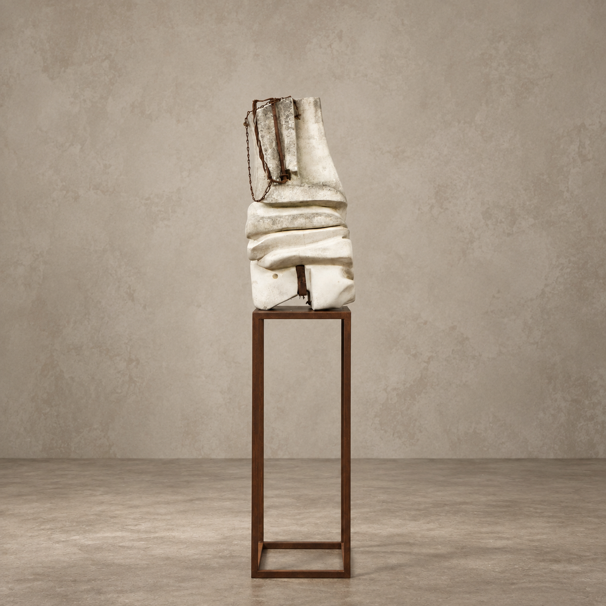A sculptural off-white draped bag with a dark chain strap, displayed on a tall dark metal pedestal against a textured beige studio wall and floor.