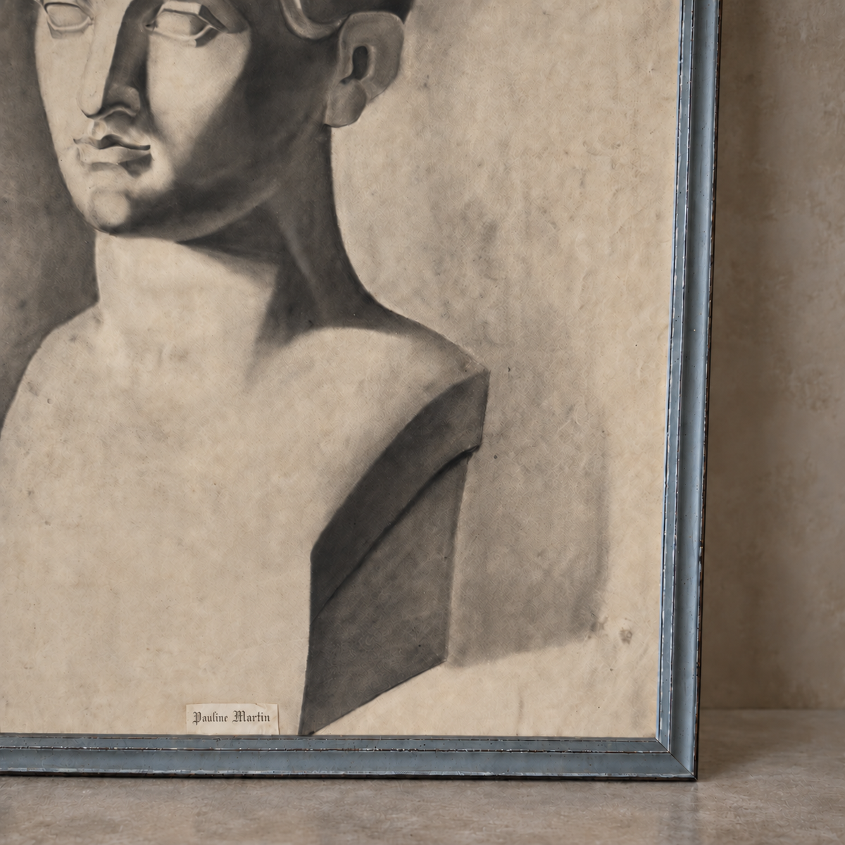 A framed monochrome artwork showing a sculptural bust with dramatic shading, photographed leaning against a wall on a floor. The frame appears dark blue-gray, and a small label near the bottom reads 'Giulio Martin'.