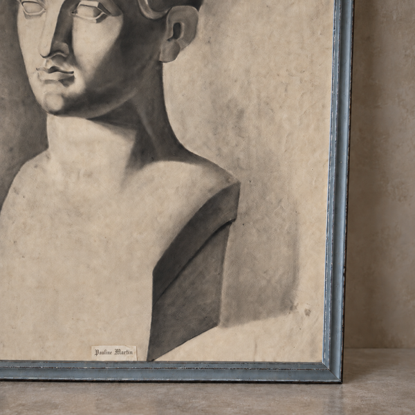 A framed monochrome artwork showing a sculptural bust with dramatic shading, photographed leaning against a wall on a floor. The frame appears dark blue-gray, and a small label near the bottom reads 'Giulio Martin'.