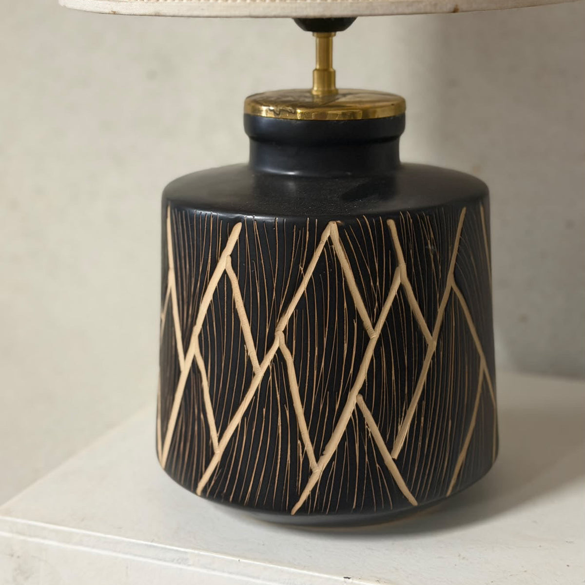 A table lamp with a glossy black ceramic base featuring a gold geometric line pattern, topped with a brass finial and a beige woven fabric lampshade.