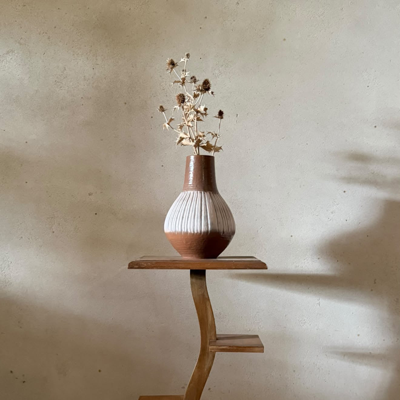 A rustic wooden display stand with three tiers holds a ribbed ceramic vase filled with dried flowers, set against a textured plaster wall with soft shadows.