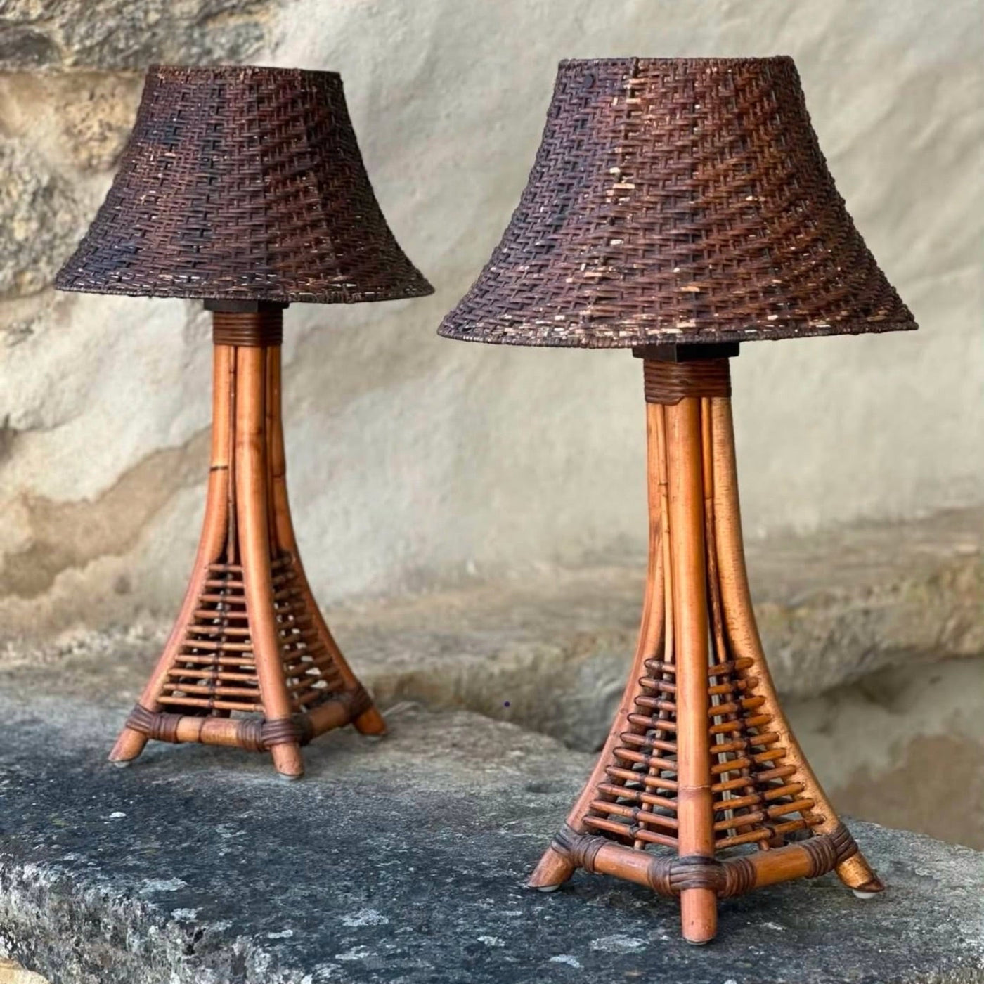 Two woven rattan table lamps with dark brown conical shades and natural bamboo-style tripod bases, displayed on a stone ledge against a stone wall. 