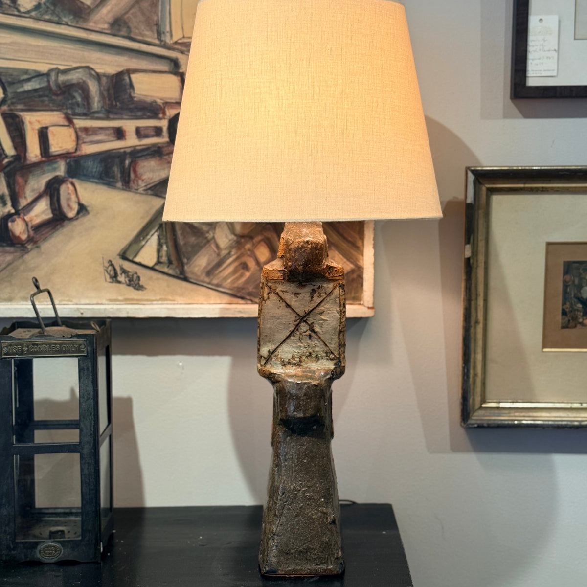 A rustic table lamp with a textured, weathered brown ceramic or stone-like sculptural base and a large beige fabric drum shade, set on a dark table against a wall with framed art.