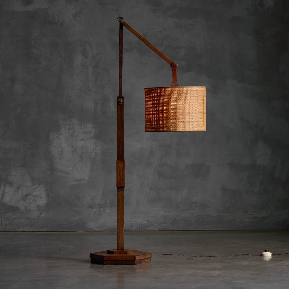 A mid-century modern floor lamp with a warm brown wooden base and arm, featuring a cylindrical woven bamboo shade, set against a textured dark gray concrete wall.