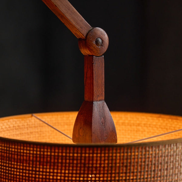Close-up of a woven cylindrical lampshade with a warm orange glow from within, supported by a dark wooden neck and arm against a dark background.