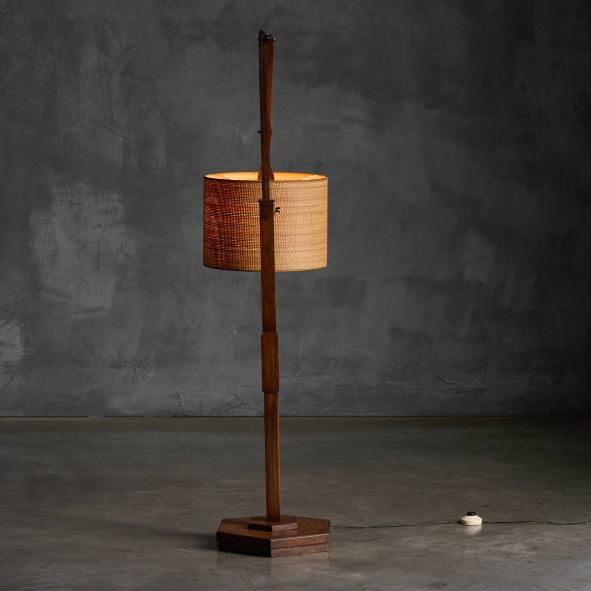 A vintage-style floor lamp with a tall wooden pole, a square wooden base, and a cylindrical woven lampshade, shown lit against a dark gray concrete wall and floor.