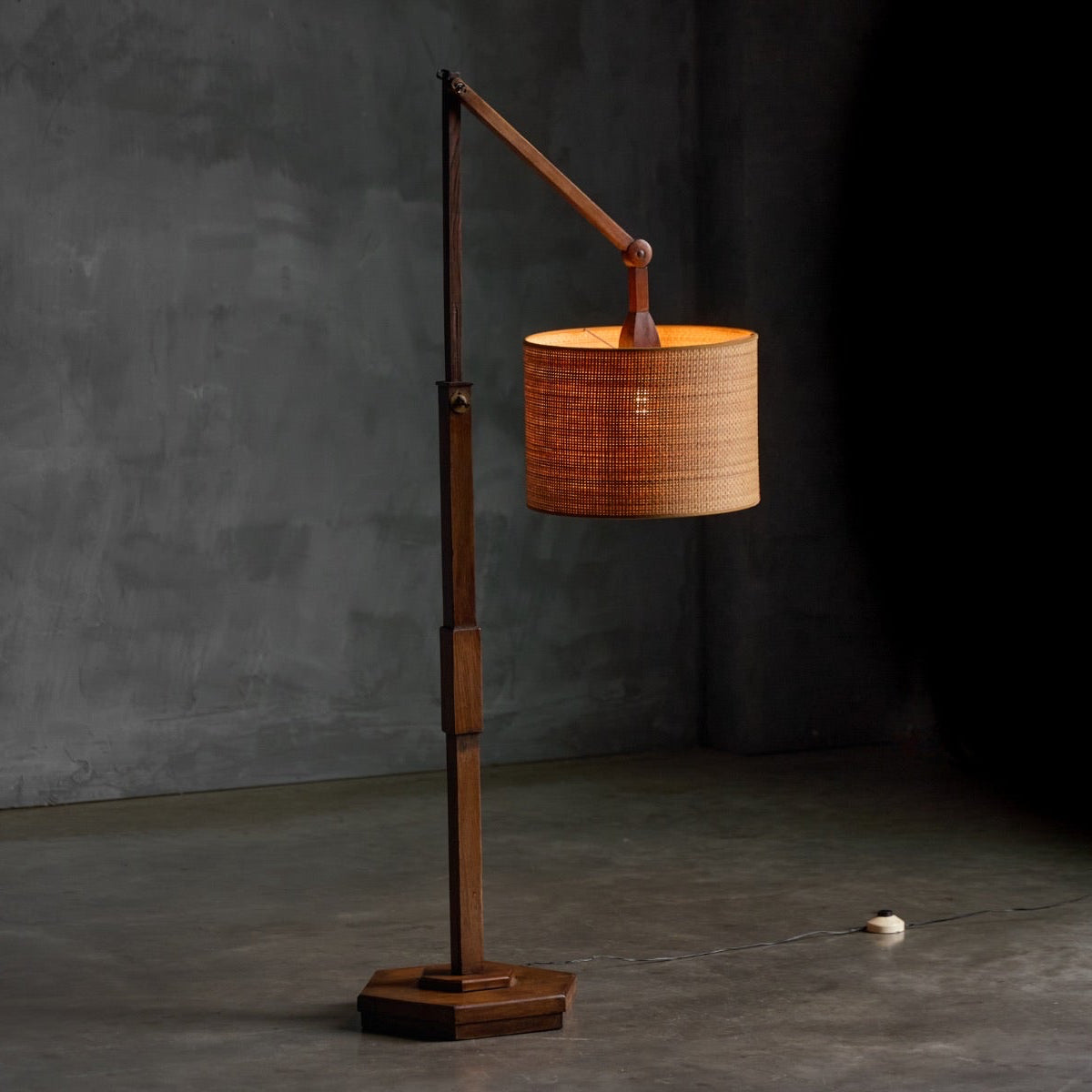 A mid-century modern floor lamp with a dark wood base and arm, featuring a cylindrical woven bamboo shade glowing warm yellow, set against a textured gray concrete wall and floor.