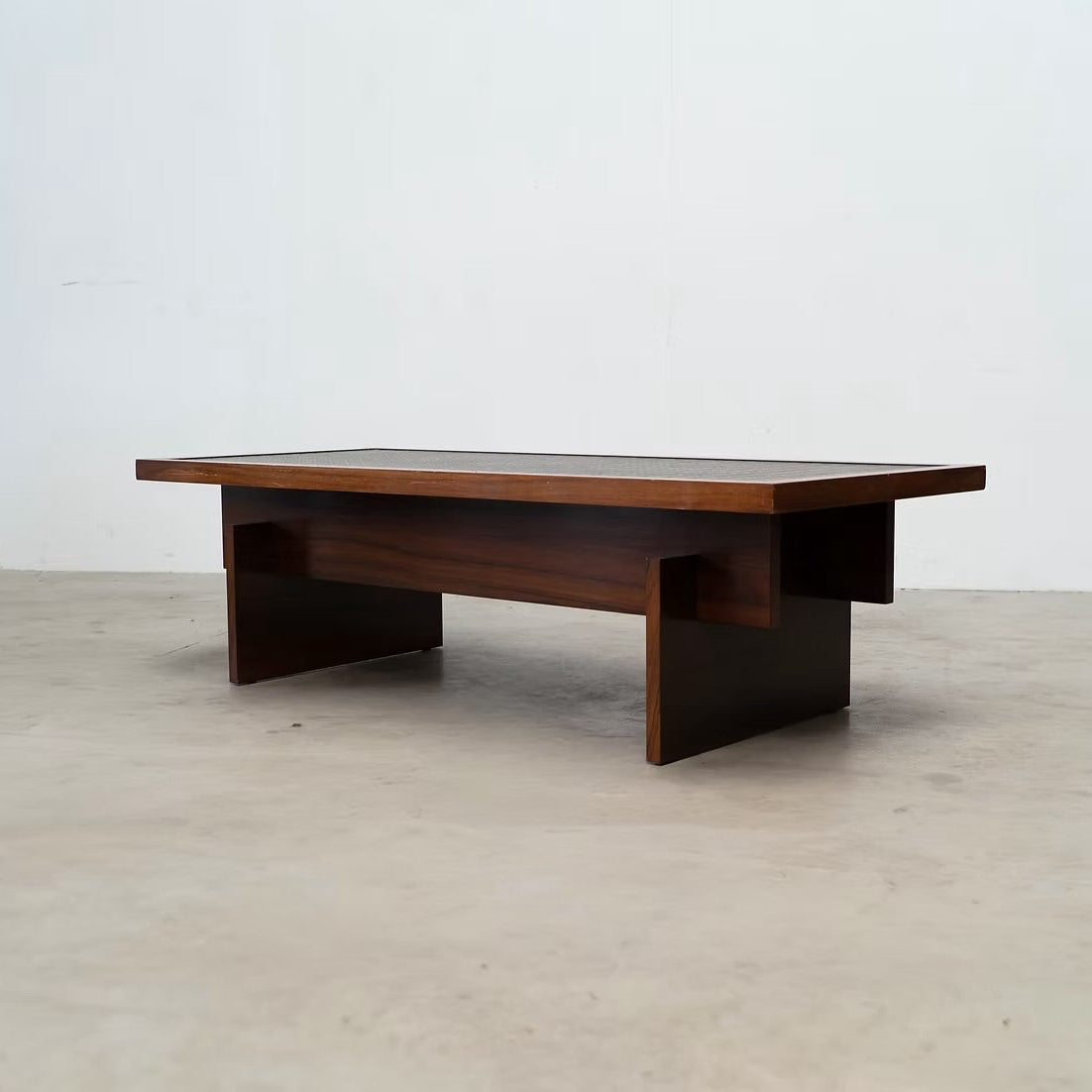 A mid-century modern wooden coffee table with a dark brown wood finish and a glass top, set against a plain white wall and concrete floor.